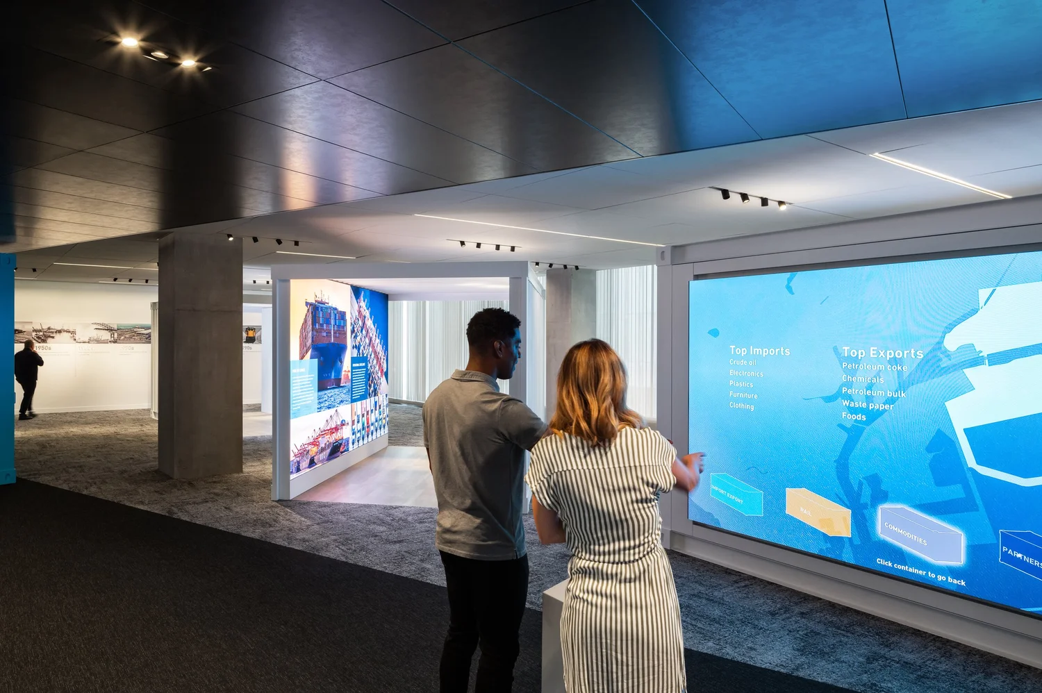 Two visitors using touch-free gestural controls on an interactive showing the Port's top imports and exports, beside a curved video tunnel of shipping imagery