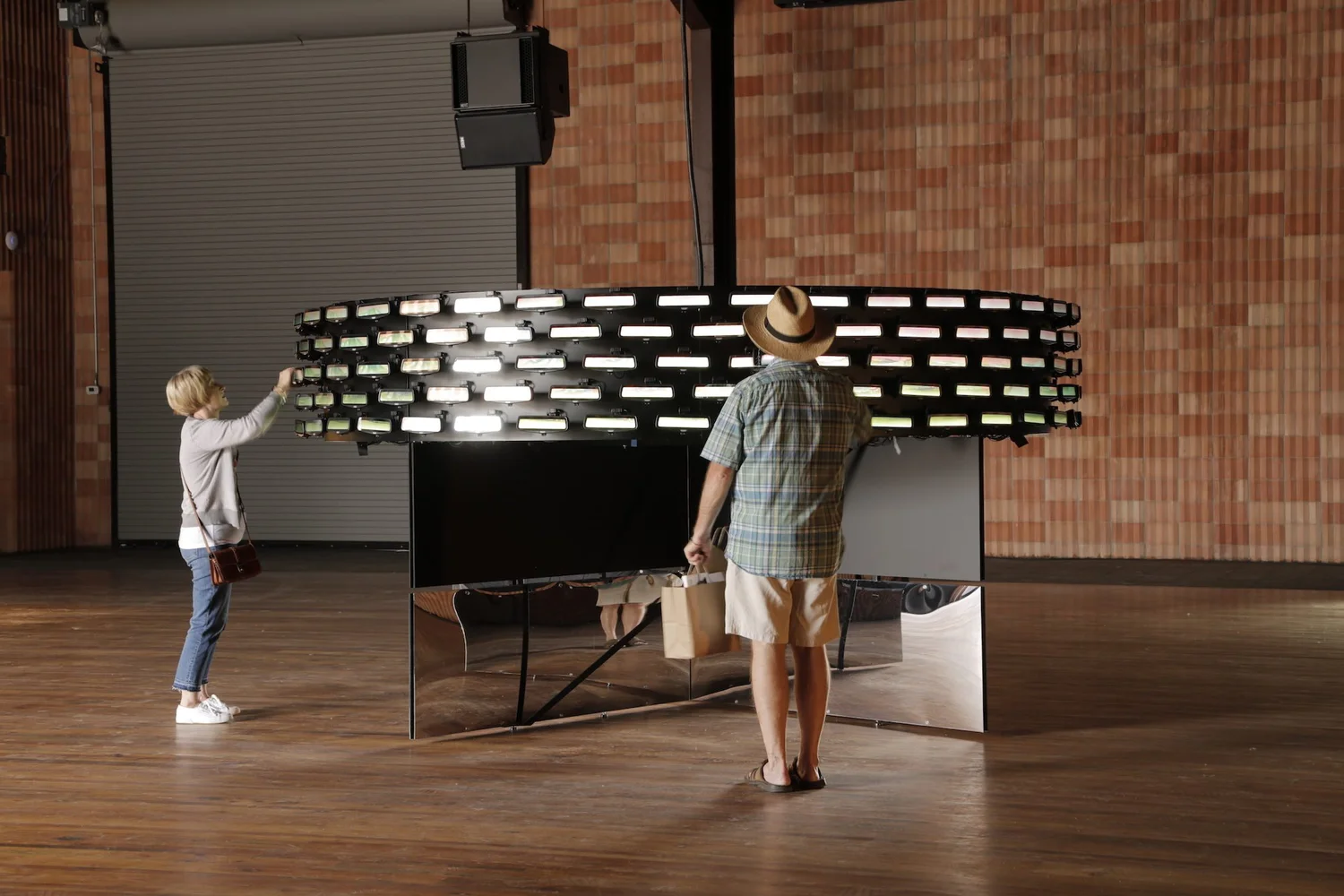 Two visitors reaching up to adjust mirrors on the suspended Horizon ring, formed from 200 Cadillac Rear Camera Mirrors, in a gallery space