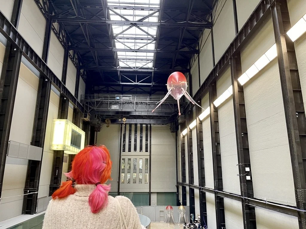 A single aerobe seen close-up from below, its tentacle-like legs trailing beneath a reflective spherical body, as a visitor watches