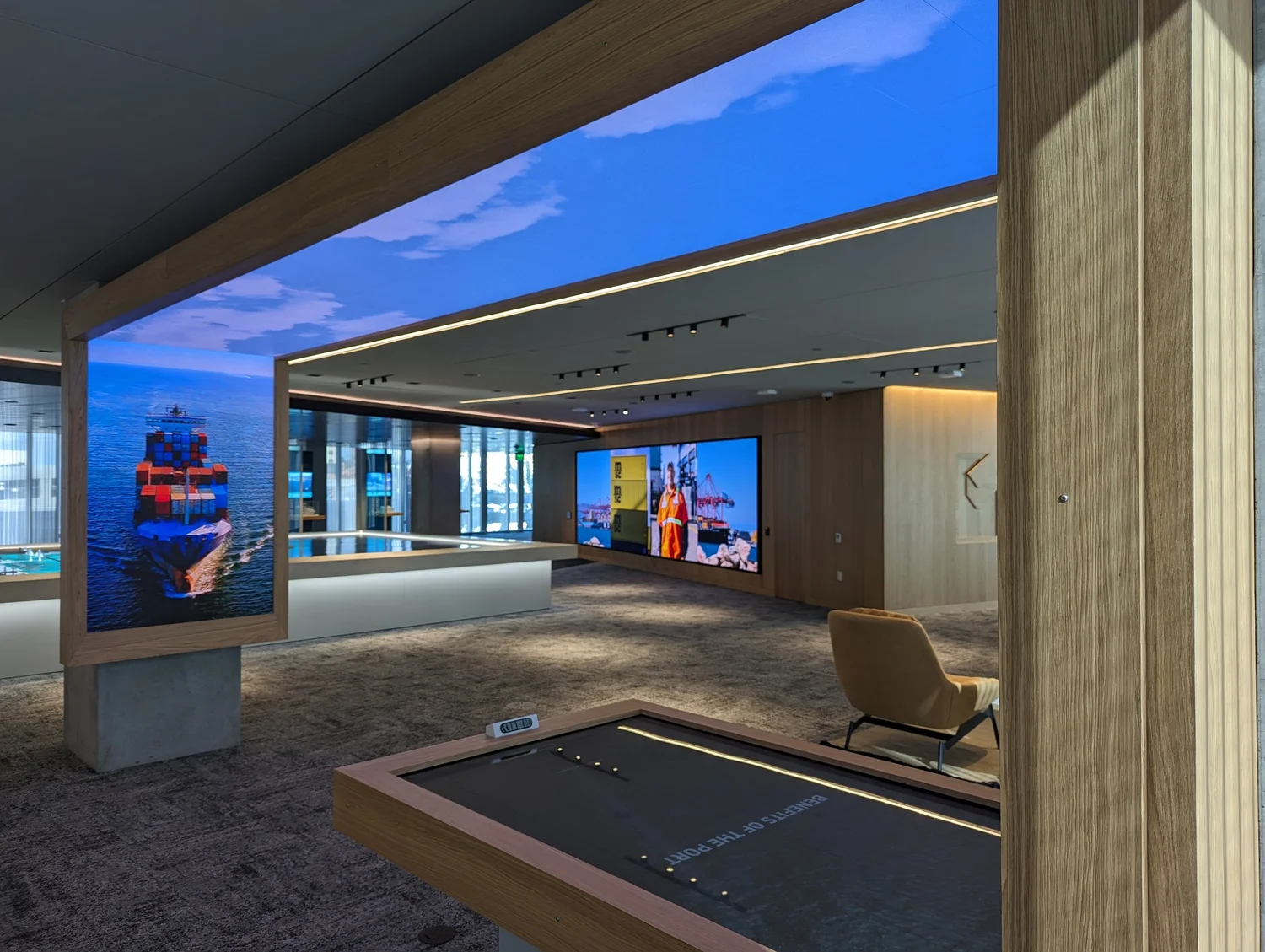 A touch-free interactive kiosk reading "Benefits of the Port" beneath a wraparound screen of a container ship, with a video wall of port operations beyond