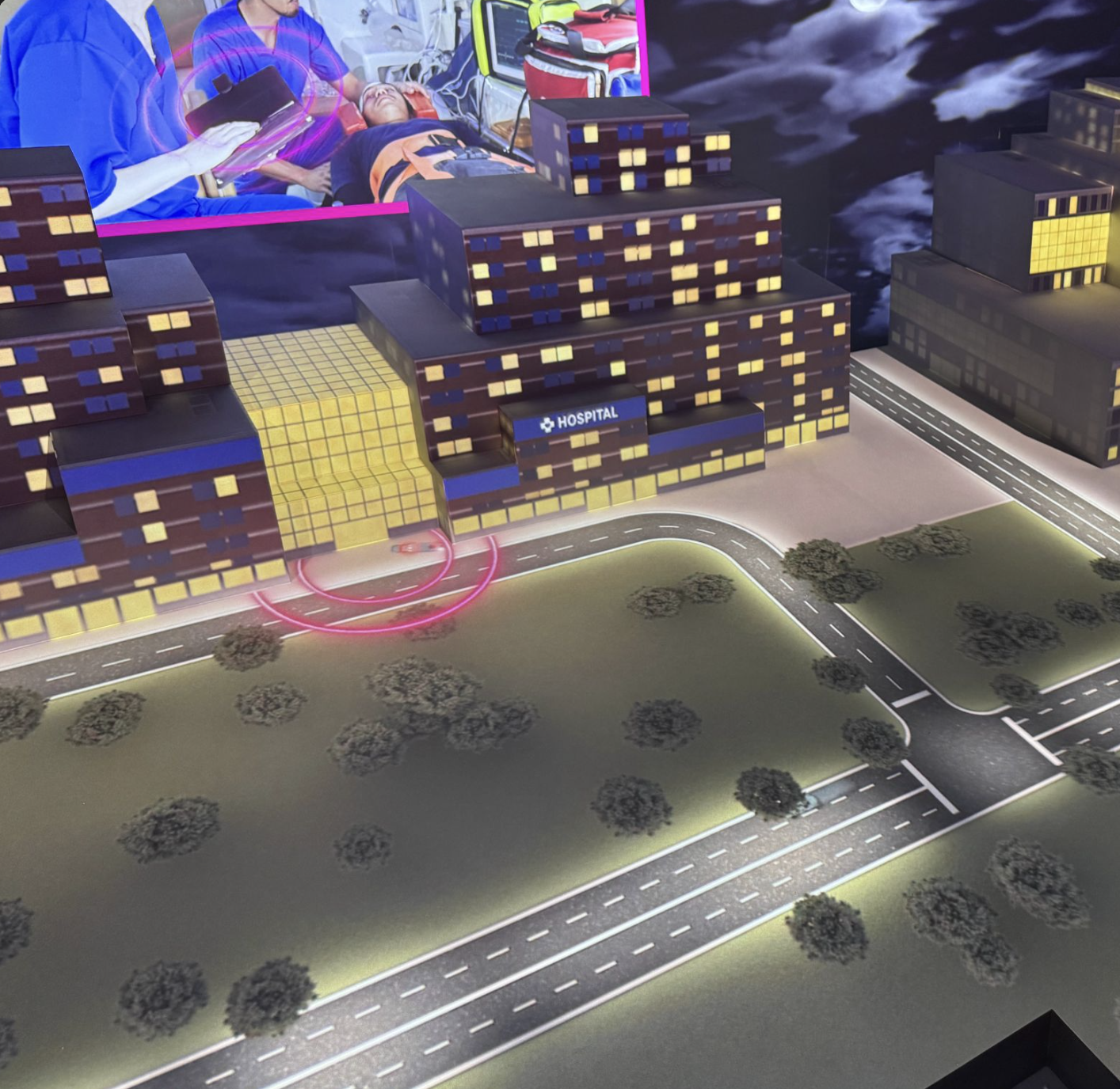 Close-up of the model's hospital building at night, with a projected inset of paramedics using connected devices and a network-coverage ring animating over the street
