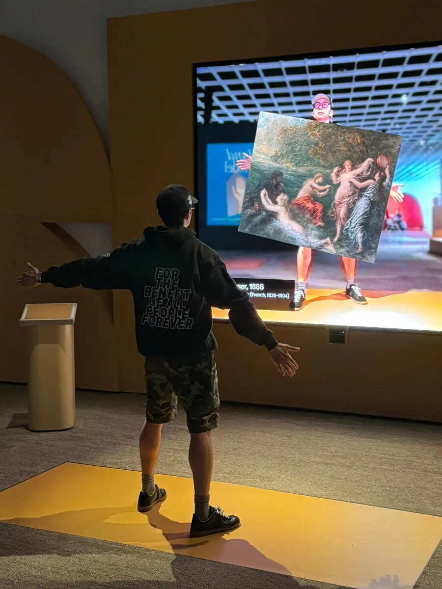 A visitor holding up the ArtLens app to compare a painting against its display on a gallery screen, part of the Art in Arm's Reach interactive