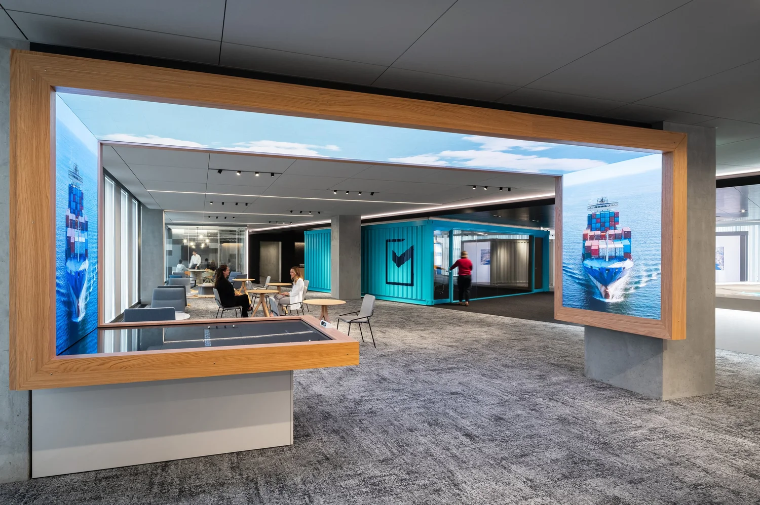 A wraparound gallery screen displaying container ships at sea, framing the center's lounge and a teal shipping-container-styled meeting room