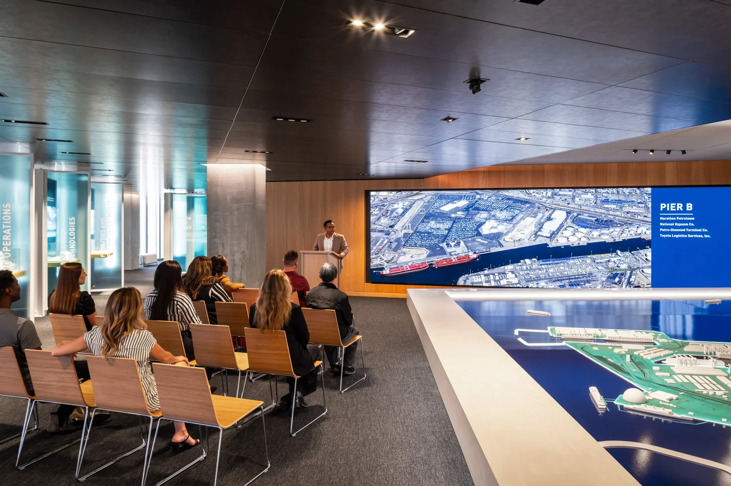 An audience in the presentation room, seated before a large aerial view of Pier B labeled with its terminal tenants, next to the illuminated harbor model